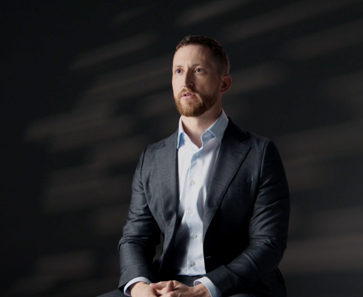 Business professional sitting on a chair, wearing a dark suit and light blue shirt, against a dark background with soft lighting, conveying a serious and contemplative expression.