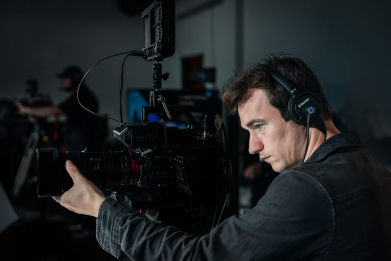 Male videographer operating a professional camera in a studio setting, focused on capturing footage, with headphones on and additional crew members visible in the background.