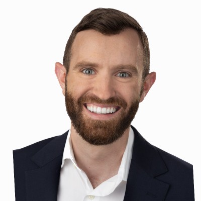 Smiling professional man with a beard and short hair, wearing a dark blazer and white shirt, against a plain white background.