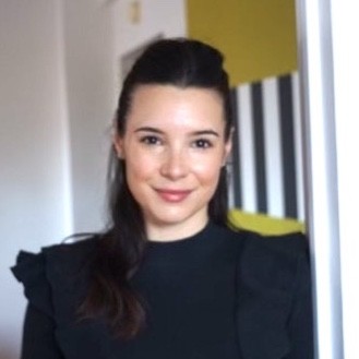 Smiling woman with dark hair and a black ruffled top, standing in a brightly lit room with a yellow accent wall and striped decor.