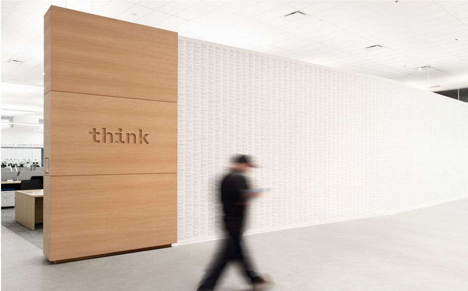 Modern office interior featuring a wooden wall with the word "think" carved into it, alongside a textured white wall. A blurred figure walks past, suggesting a dynamic workspace environment.