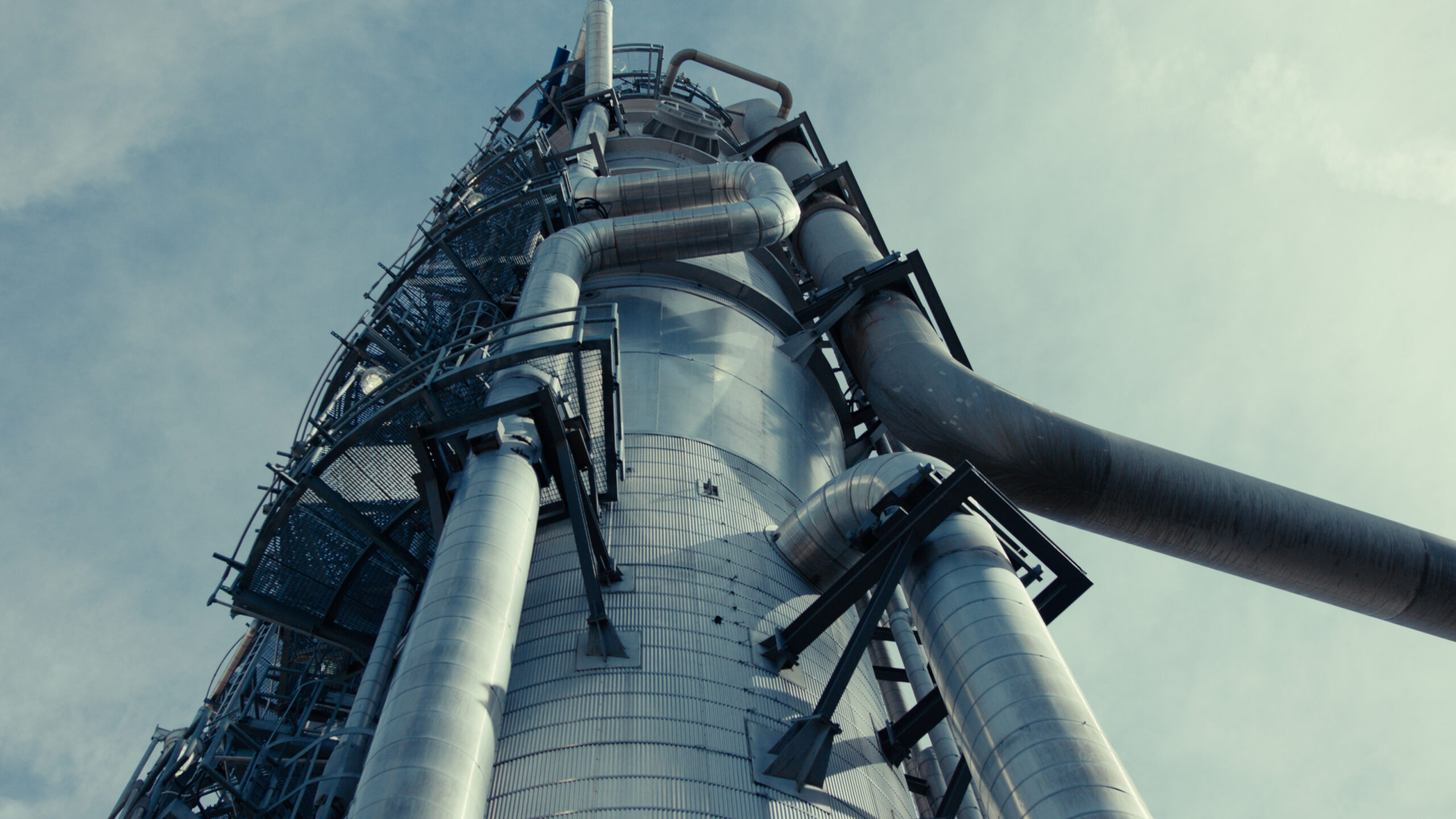 Industrial smokestack with metallic pipes and structures, viewed from below against a cloudy sky. The image showcases modern engineering and design in an industrial setting, highlighting the complex arrangement of pipes and framework.