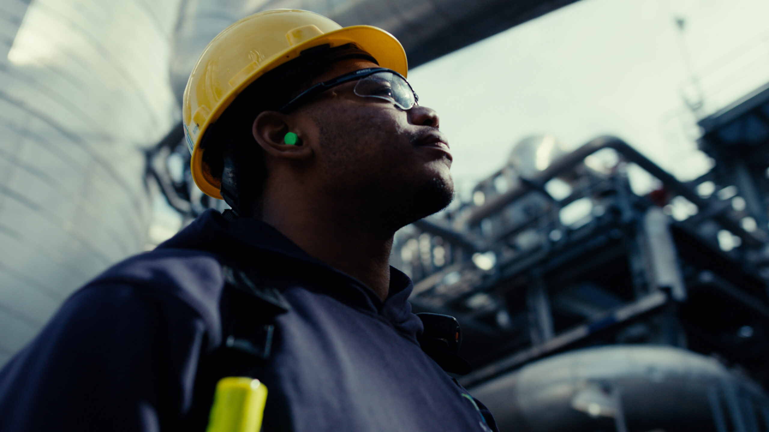 Construction worker wearing a yellow hard hat and safety glasses, looking up thoughtfully at an industrial site with machinery in the background.