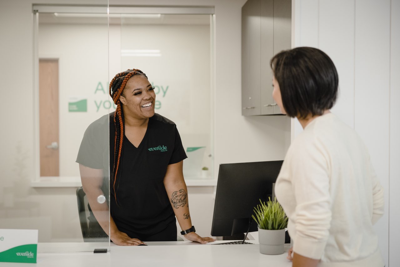 Smiling healthcare professional at reception desk engaging with a patient in a modern medical office, showcasing friendly service and community-focused care.