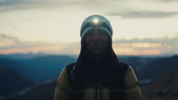 A hiker wearing a headlamp and a yellow jacket stands against a scenic mountain backdrop at dusk, showcasing outdoor adventure and exploration.