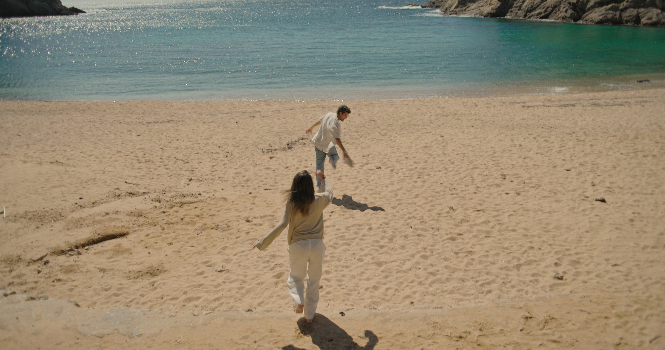 A couple enjoys a playful moment on a sandy beach, with turquoise waters in the background and rocky cliffs in the distance, capturing the essence of a sunny day by the ocean.