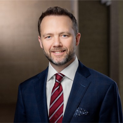 Professional headshot of a man in a dark suit and red striped tie, smiling confidently against a neutral background.
