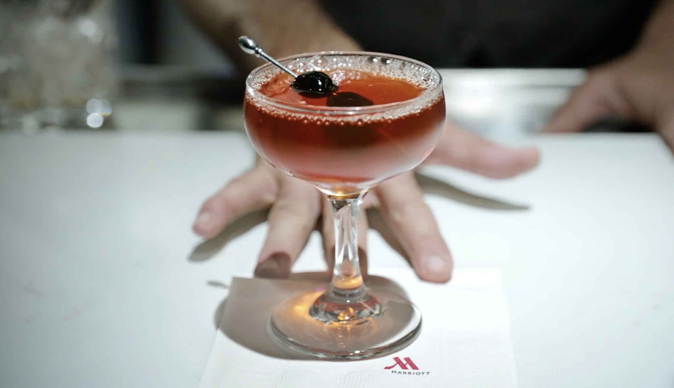 Cocktail served in a classic coupe glass, featuring a vibrant red color and garnished with a cherry. The drink is placed on a white napkin with the Marriott logo, with a bartender's hands partially visible in the background.