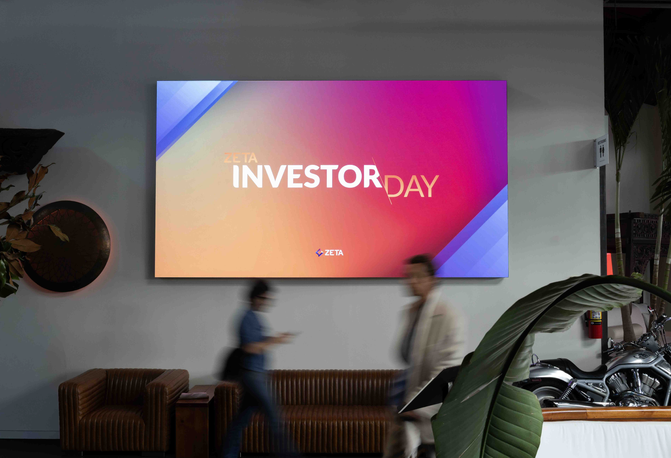 Zeta Investor Day event display featuring a vibrant digital screen with the text "INVESTOR DAY" in a modern font, set against a colorful background. Two blurred figures are walking past a stylish brown couch and decorative elements, including a motorcycle and plants in the foreground.