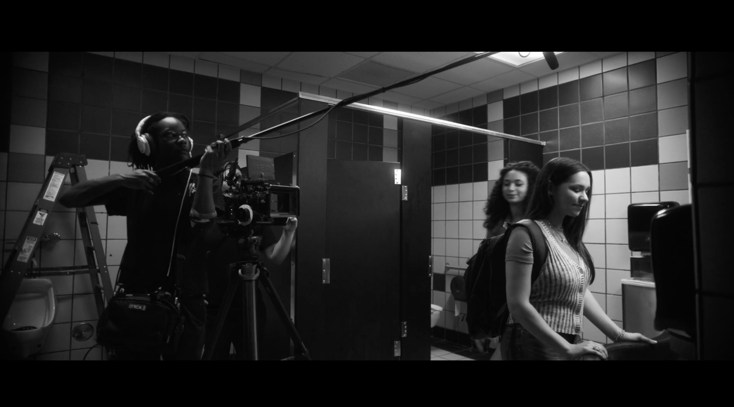 Behind-the-scenes image of a film crew in a school bathroom, featuring a camera operator with a boom mic, a cinematographer adjusting equipment, and two female actors preparing for a scene. The setting is characterized by colorful tiled walls and typical bathroom fixtures.