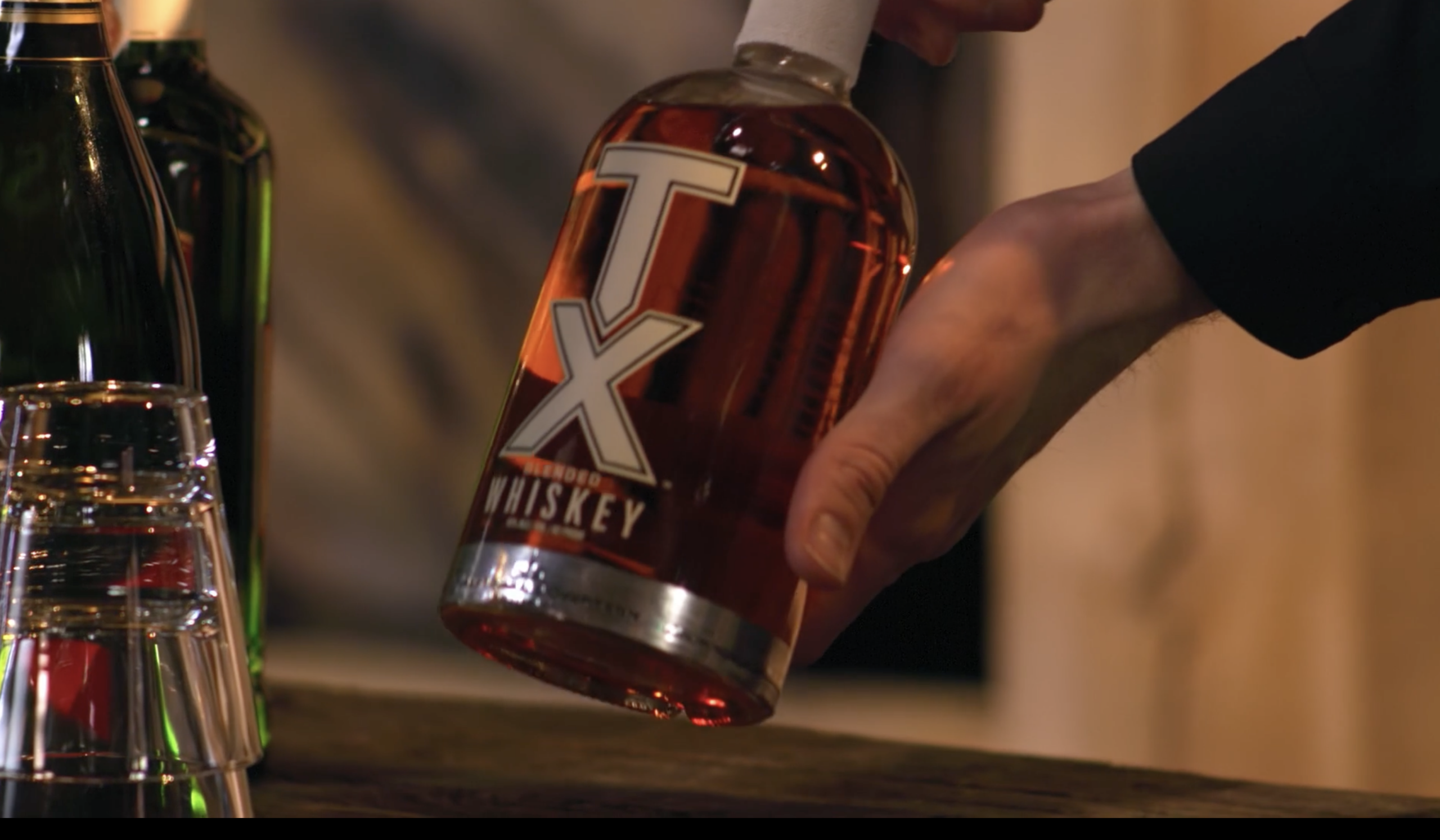 A hand holding a bottle of TX Whiskey, showcasing its amber color and distinctive label, with glassware in the foreground and other liquor bottles blurred in the background.