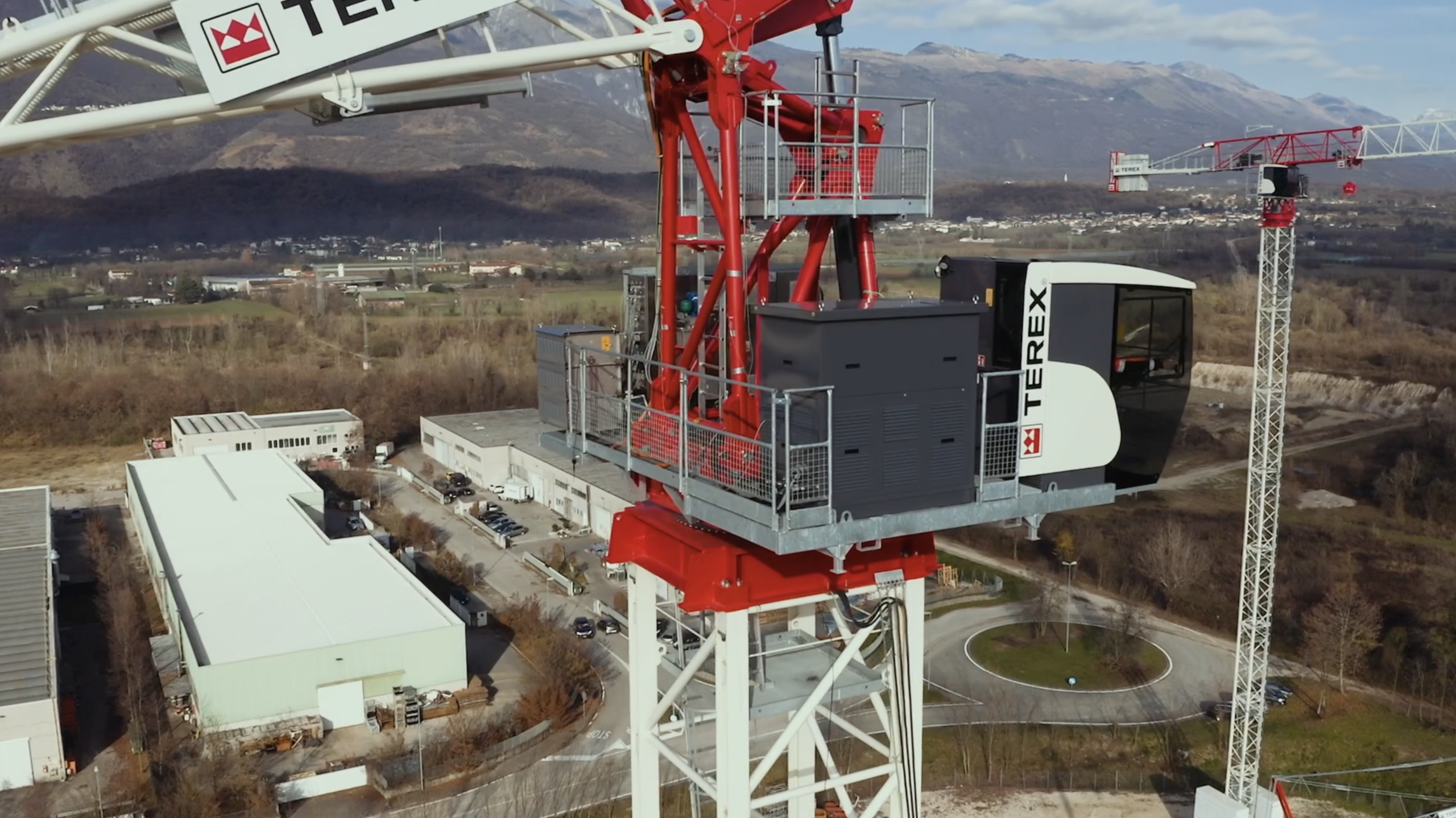 Aerial view of a Terex construction crane with a cabin, showcasing its red and white structure against a mountainous backdrop and industrial landscape.