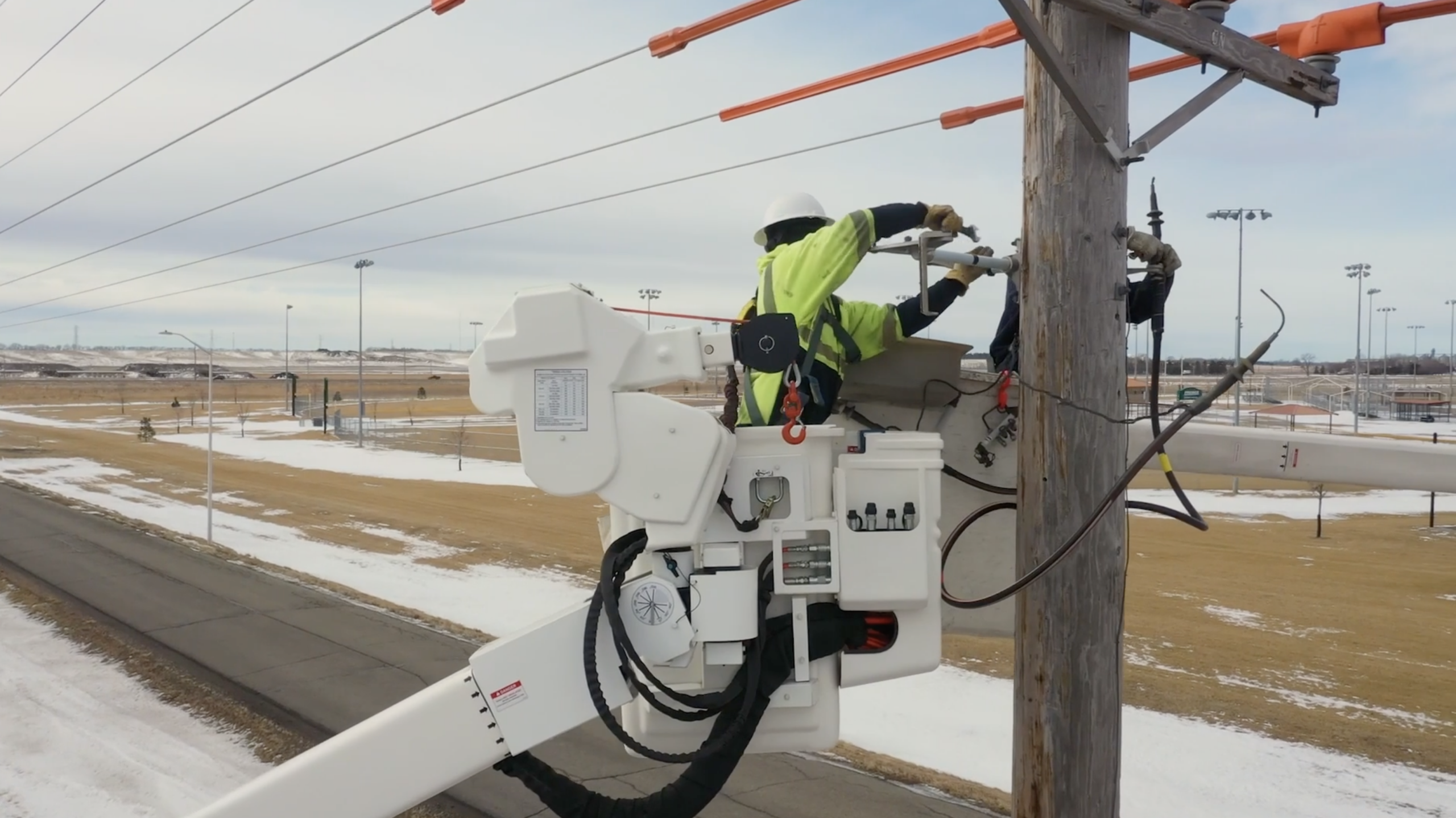 Utility worker performing maintenance on power lines from a bucket truck, with a focus on safety gear and equipment, in a snowy landscape.