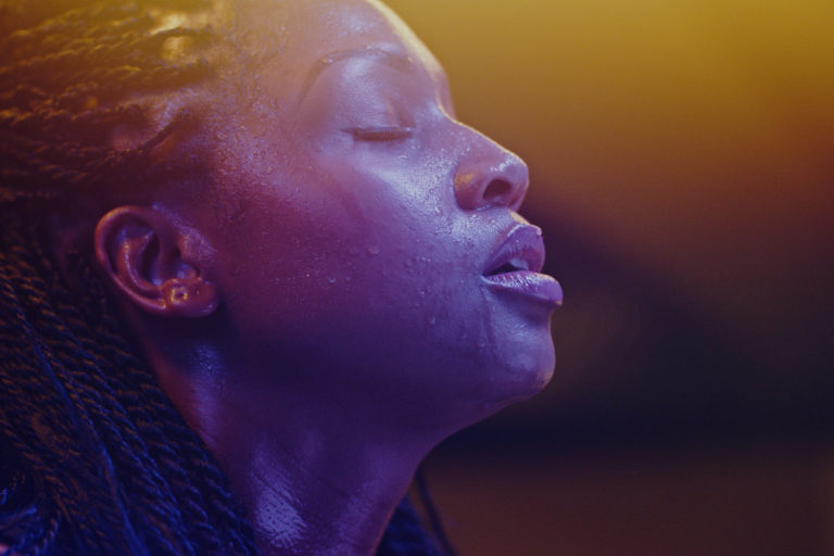 Close-up of a woman with braided hair, eyes closed and face glistening with sweat, illuminated by soft, colorful lighting, conveying a sense of tranquility and intensity.