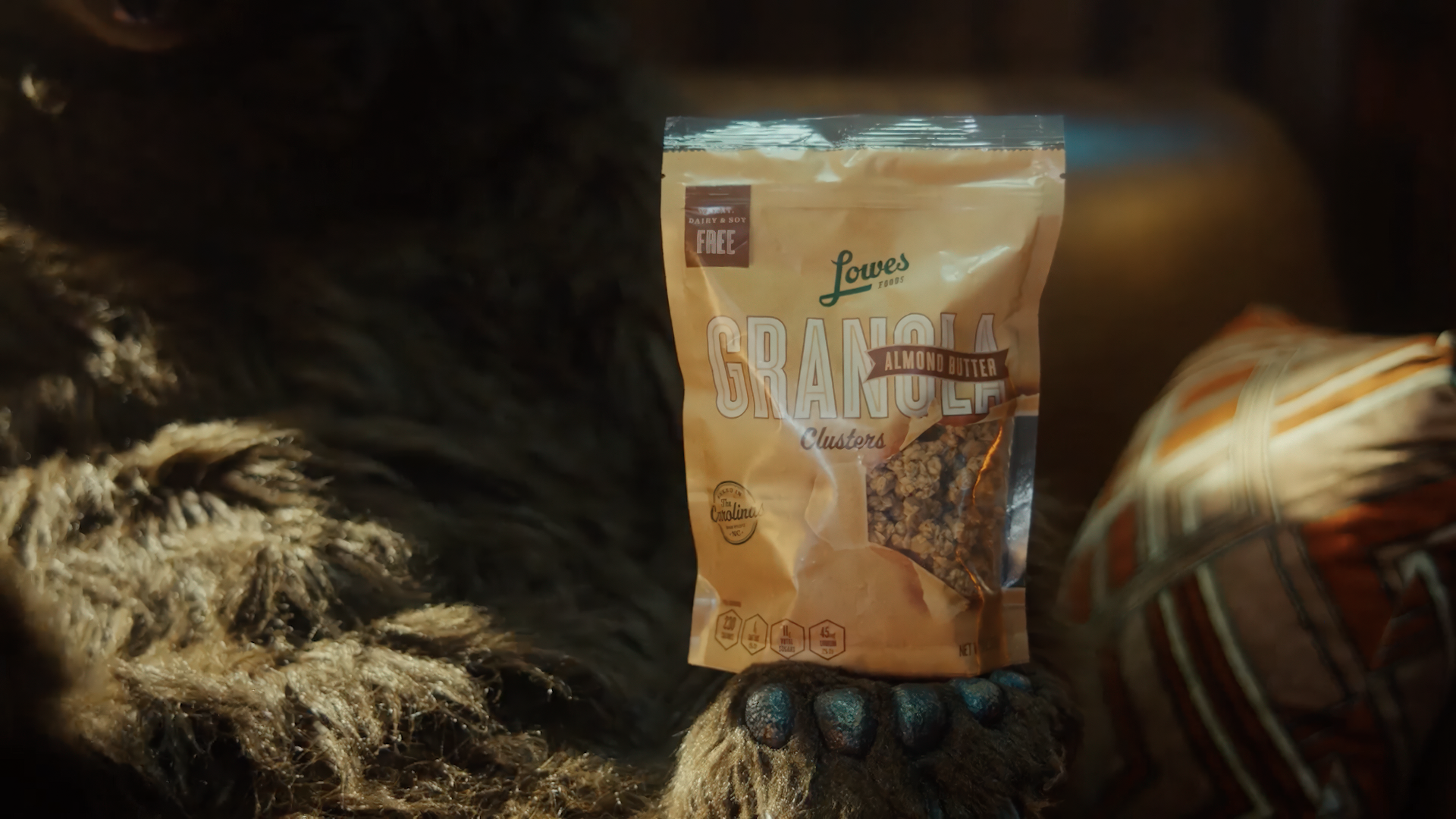 Granola clusters in an almond butter flavor, packaged in a Lowes Foods bag, held by a furry creature, with a cozy background featuring a patterned pillow. Ideal for healthy snacking, dairy-free, and soy-free.