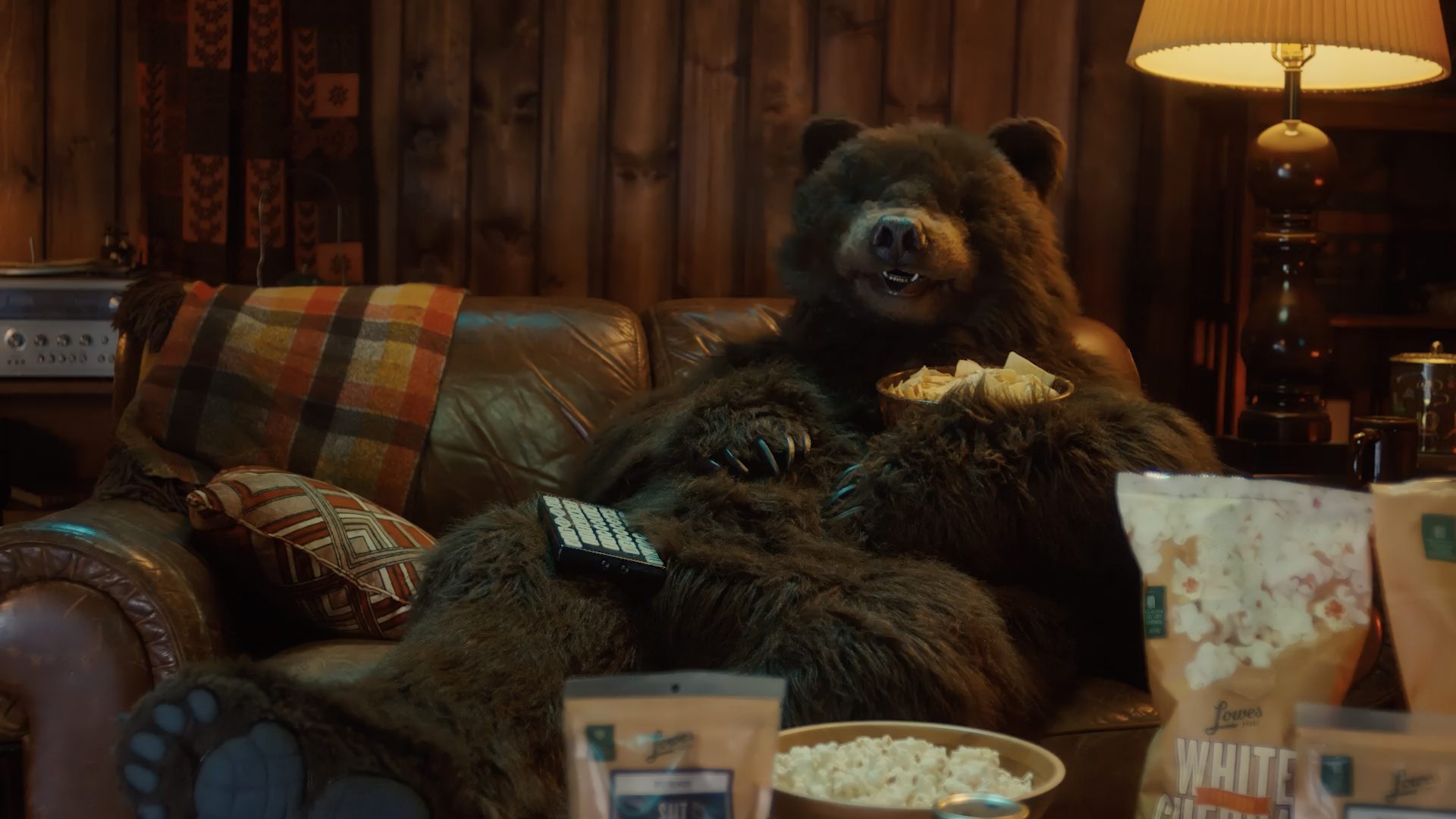A cozy living room scene featuring a bear lounging on a leather couch, enjoying snacks from a bowl while holding a remote control. Surrounding the bear are bowls of popcorn and various snack bags, with warm wooden paneling in the background and a lamp providing soft lighting.