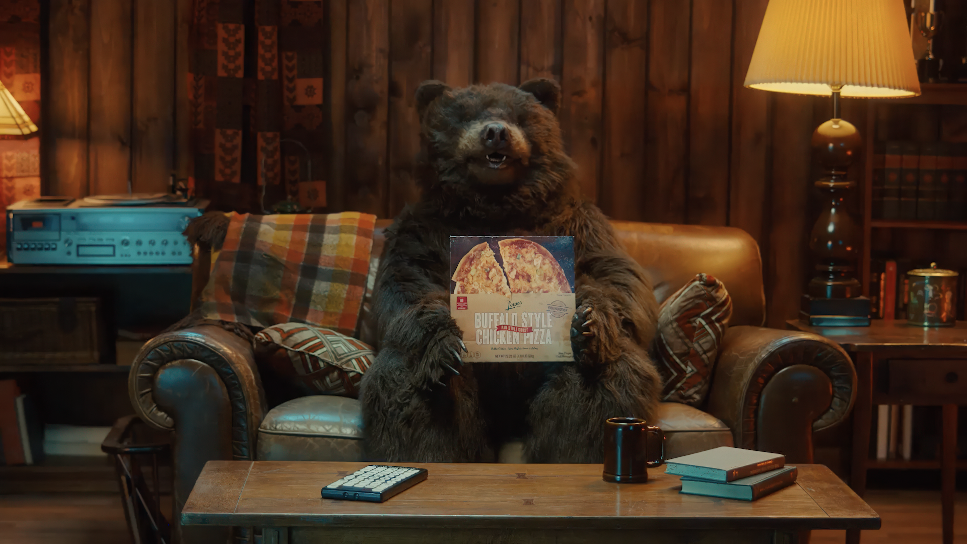 A bear sitting on a leather couch in a cozy, rustic living room, holding a box of Buffalo Style Chicken Pizza, with a coffee mug and a remote control on the table.
