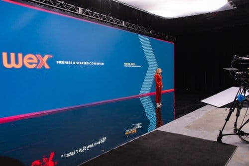 Presenter in a red suit delivering a business and strategic overview for Wex on a modern stage with a blue backdrop and professional lighting setup. Camera equipment is visible in the foreground, indicating a live event or presentation setting.