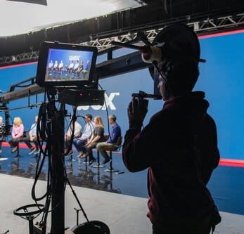 A camera operator using a crane to film a panel discussion in a studio setting, with a blue backdrop and a group of speakers seated on stage.
