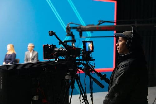 Camera operator capturing a video production scene with two presenters in the background, featuring a blue backdrop and modern lighting setup.