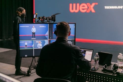 A production team setting up for a live event, featuring a person operating a control panel with multiple screens displaying a presenter on stage, while another crew member prepares equipment in the background. The backdrop displays the Wex logo, emphasizing technology and event production.