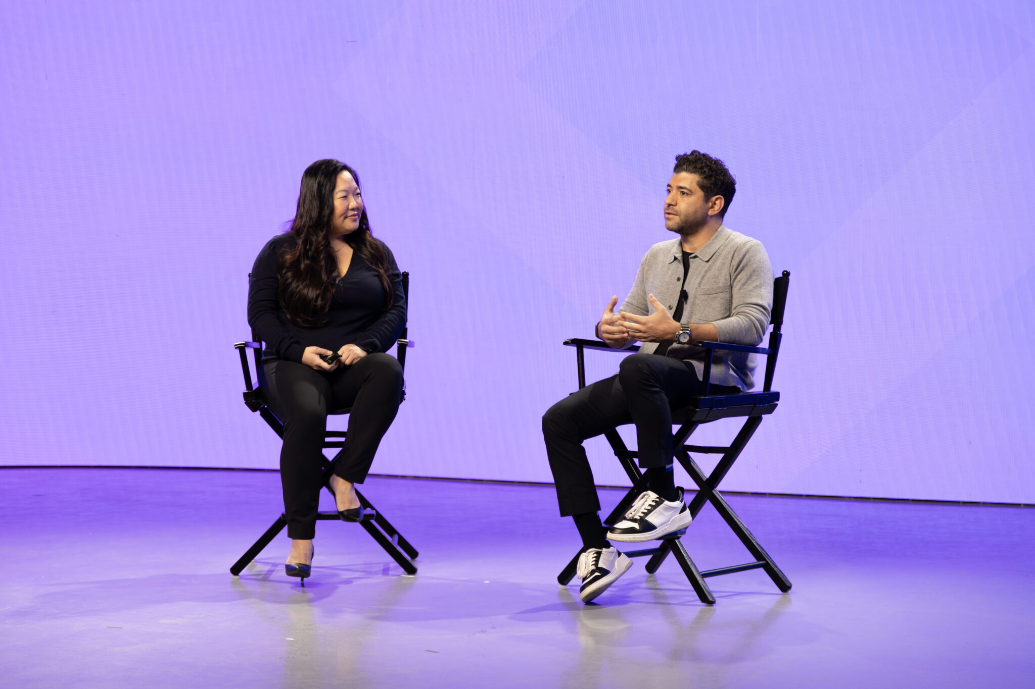 Two speakers engaged in a discussion on stage, seated in director's chairs against a vibrant purple backdrop. The woman on the left has long hair and is dressed in a black top and pants, while the man on the right is wearing a gray sweater and black pants, gesturing as he speaks. The setting appears to be a professional event or conference focused on dialogue and insights.