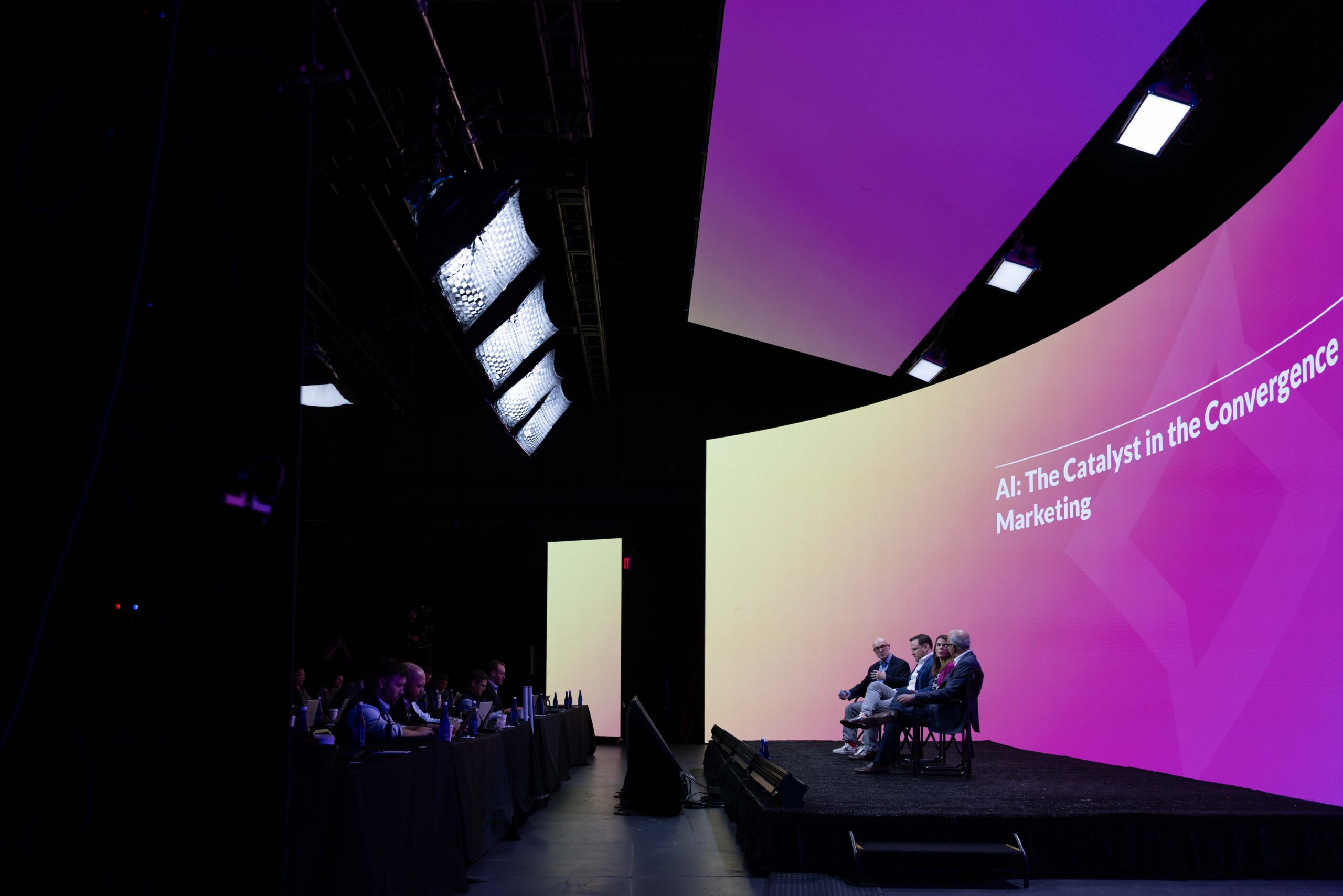 Panel discussion on AI in marketing, featuring speakers seated on stage with a colorful backdrop and audience members engaged at tables. The topic highlighted is "AI: The Catalyst in the Convergence Marketing."