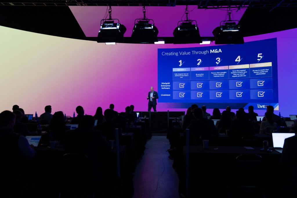 Business conference presentation on creating value through M&A, featuring a speaker in front of a large screen displaying criteria for successful mergers and acquisitions, with an audience engaged in the background.