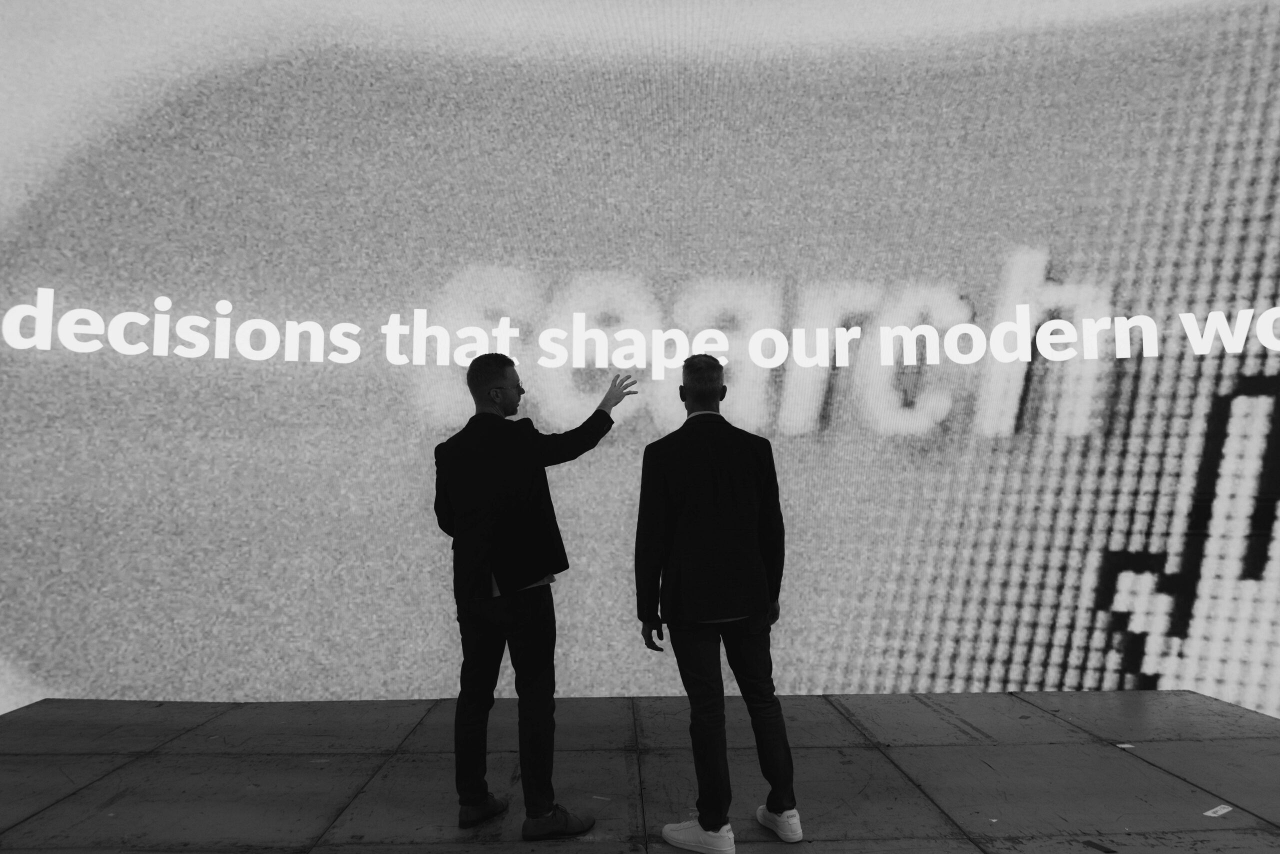 Two individuals engaged in a discussion in front of a large screen displaying the phrase "decisions that shape our modern world," highlighting themes of technology and decision-making.