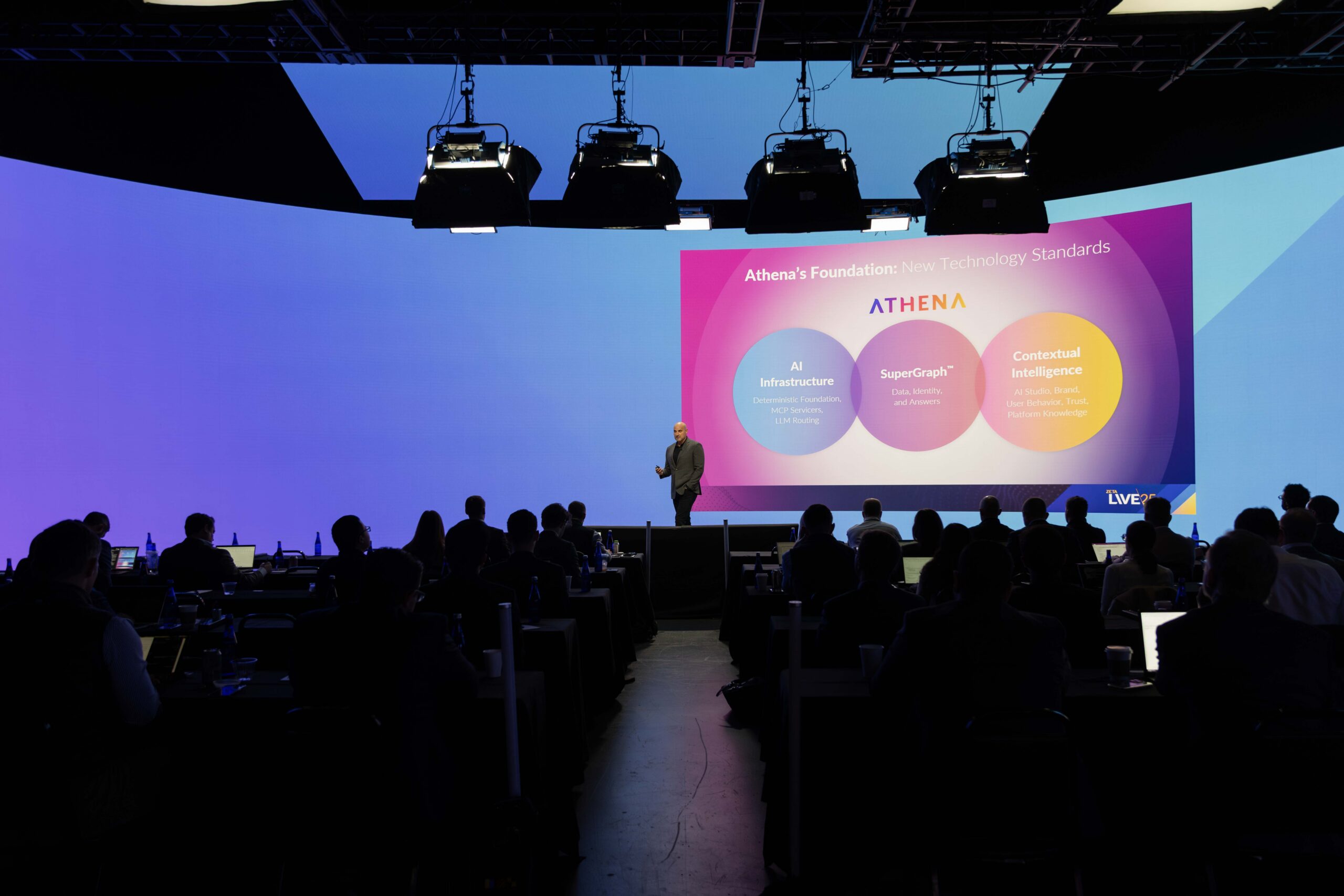 A speaker presents on stage at a technology conference, discussing Athena's foundational technology standards, including AI Infrastructure, SuperGraph, and Contextual Intelligence, with an audience engaged in the background.