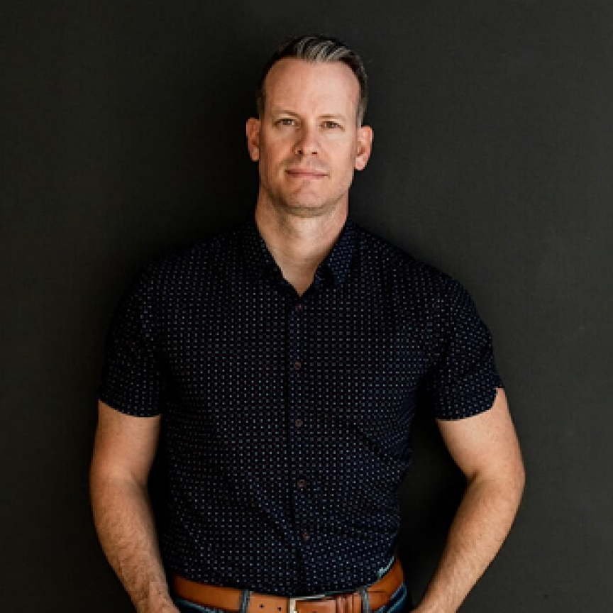 Portrait of a man with short hair, wearing a dark patterned shirt and standing against a black background, conveying a confident and approachable demeanor.