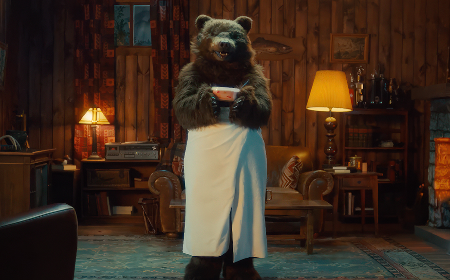 A bear dressed in a white apron stands in a cozy cabin living room, holding a bowl with a spoon. The room features wooden walls, a lamp, and a vintage record player, creating a warm and inviting atmosphere.