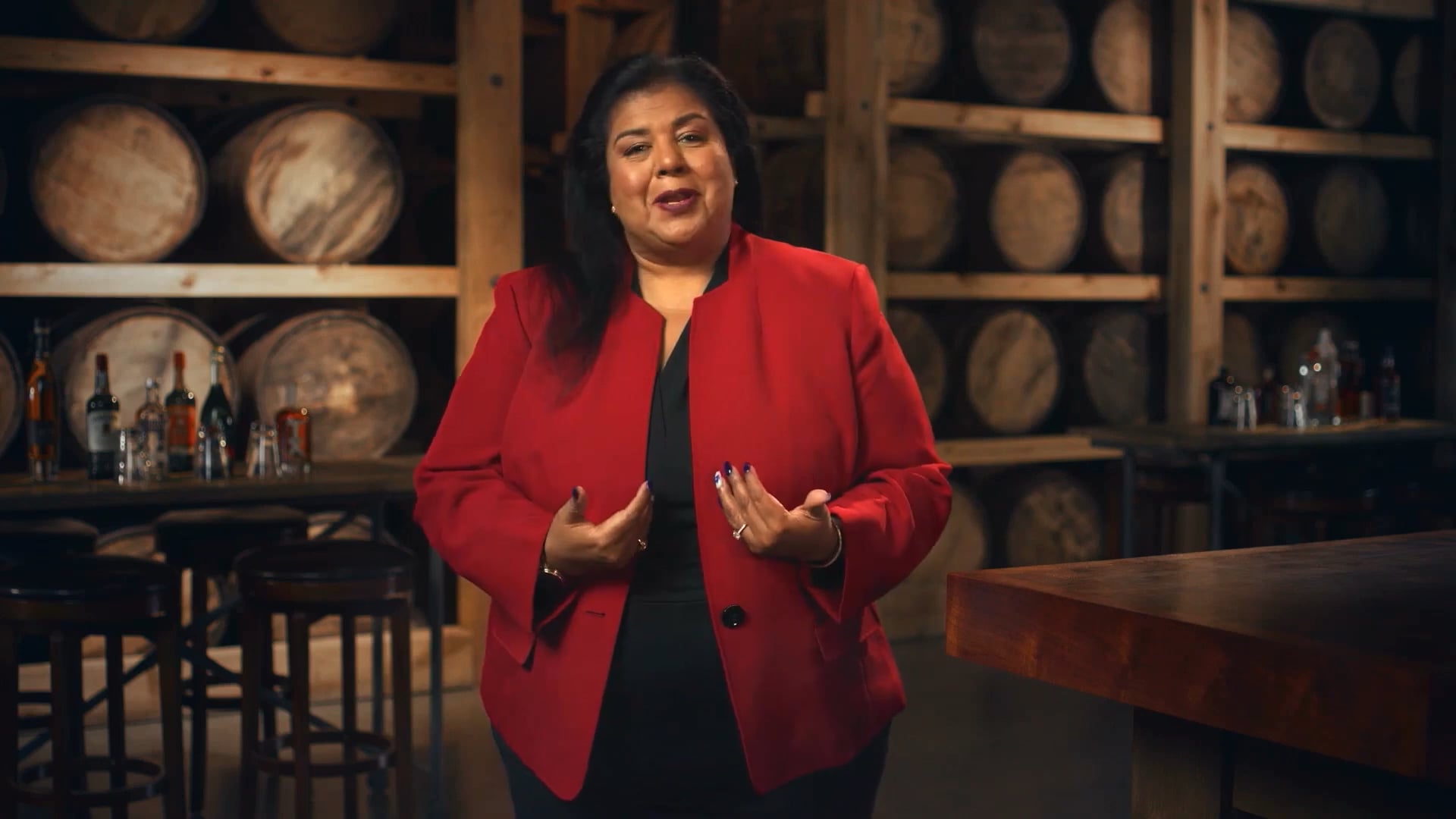 Woman in a red blazer speaking confidently in a distillery setting, with wooden barrels and various bottles in the background. The scene conveys expertise in spirits and hospitality, ideal for content related to beverage industry presentations or interviews.